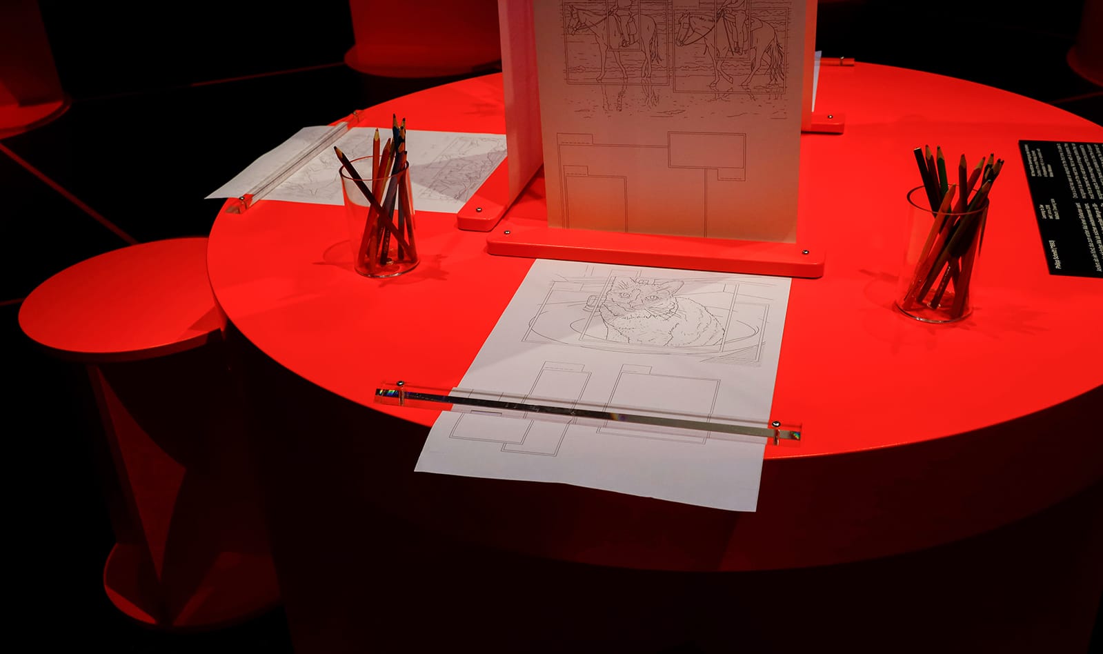 A red table with three seats, each with a roll of coloring pages in front of them. It is an art exhibit at Deutsches Hygiene Museum Dresden