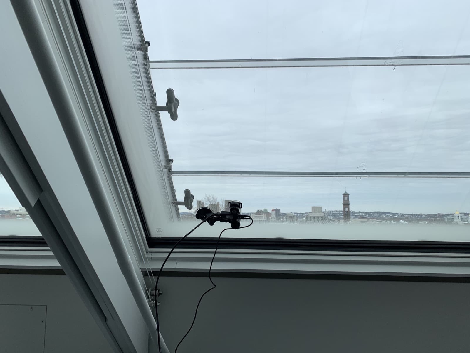 A photo of a small camera attached to a glass ceiling. Through the glass a skyline with a curchtower is visible. The sky is overcast in a pale, gray blue.